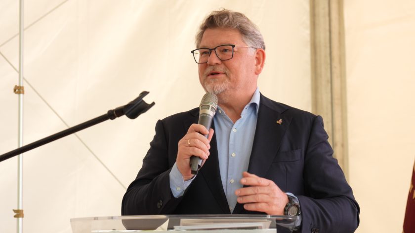 Zbliżenie na mężczyznę w garniturze trzymającego mikrofon podczas przemówienia. W tle biały namiot konferencyjny.