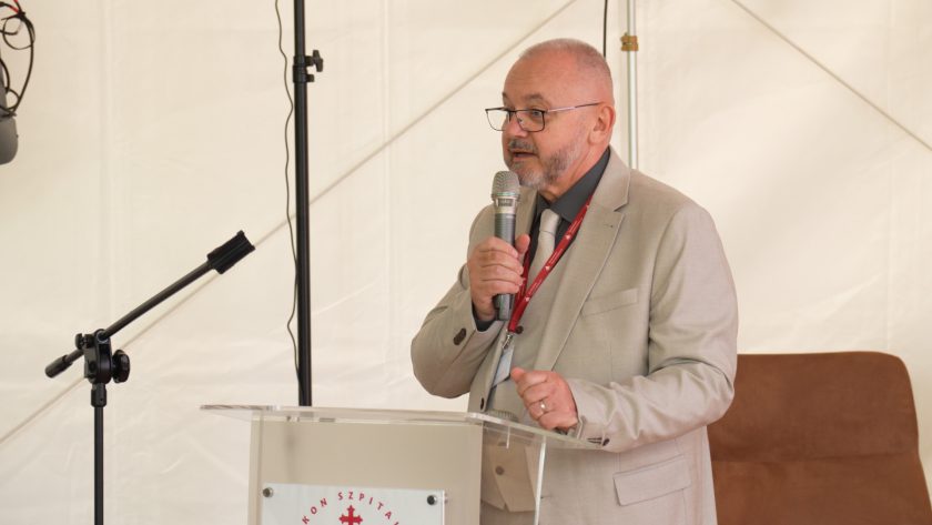 Mężczyzna w jasnym garniturze przemawia z mównicy. W tle biały namiot i elementy nagłośnienia.