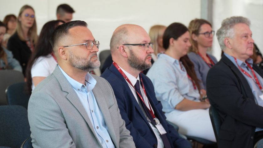 Dwóch mężczyzn w garniturach siedzi wśród publiczności, uważnie słuchając prezentacji. W tle pozostali uczestnicy konferencji.