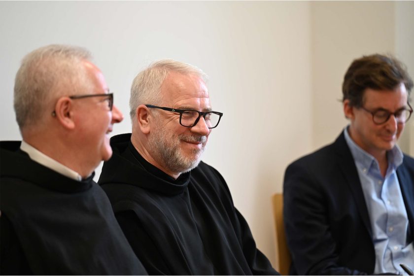 Dwóch zakonników w czarnych habitach uśmiecha się podczas rozmowy. Obok nich mężczyzna w garniturze patrzy w dół, jakby czytał lub pisał.