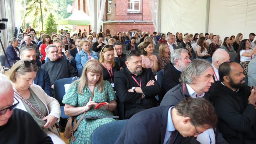 ełna sala uczestników konferencji siedzących pod białym namiotem. Widzowie słuchają uważnie prezentacji, w tle widoczny czerwony budynek i zielone namioty.