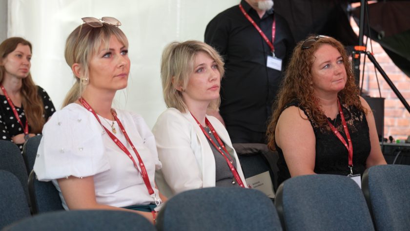 Trzy kobiety siedzące w pierwszym rzędzie w skupieniu słuchają wystąpienia; wszystkie mają zawieszone na szyi identyfikatory na czerwonych smyczach. W tle widoczna jest kobieta stojąca i osoby siedzące w dalszych rzędach.