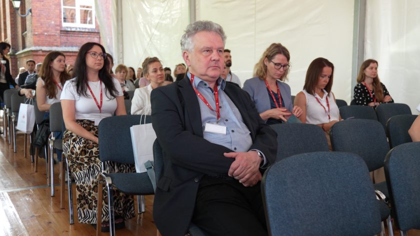 Uczestnicy konferencji zajmujący tylne rzędy krzeseł; mężczyzna na pierwszym planie siedzi ze skrzyżowanymi rękami, obok niego młoda kobieta w białej bluzce. W tle widoczne kolejne osoby, niektóre przeglądają telefony.