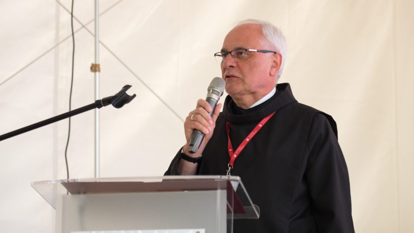 Brat zakonny mówi do mikrofonu, stojąc przy mównicy z logo Bonifraterskiego Centrum Medycznego; ubrany jest w czarny habit, a w tle widoczny jest fragment białej ścianki namiotu.