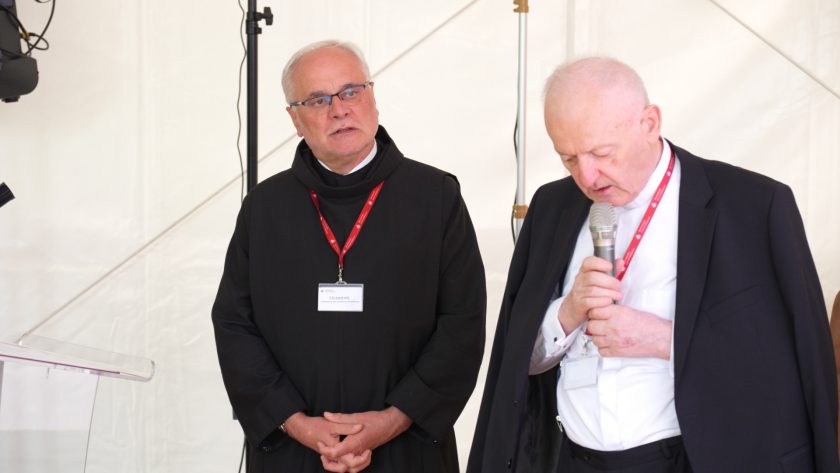 Brat zakonny i duchowny w garniturze stoją razem przy mównicy; zakonnik z założonymi rękami słucha, podczas gdy drugi mężczyzna modli się ze spuszczoną głową i mikrofonem w dłoni.