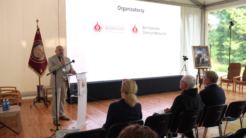 Mężczyzna w beżowym garniturze wygłasza przemówienie przy mównicy, na ekranie w tle wyświetlone są logotypy organizatorów wydarzenia. Przed nim siedzą słuchacze – również widoczny zakonnik i portret św. Jana Bożego na sztaludze.
