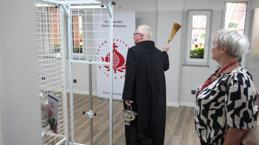 Brat zakonny w habicie błogosławi pomieszczenie za pomocą kropidła; obok niego stoi kobieta z identyfikatorem, w tle baner Bonifraterskiego Centrum Medycznego oraz okna z doniczkami pelargonii.