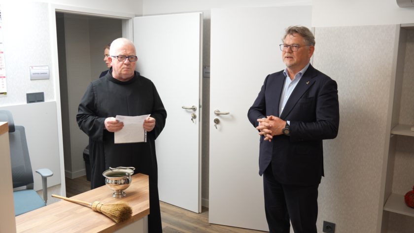 Brat zakonny trzymający kartkę papieru oraz mężczyzna w garniturze stoją razem w jednym z nowych pomieszczeń szpitala. Na blacie przed nimi leży kropidło i naczynie z wodą święconą. W tle drzwi oznaczone jako „szatnia damska” i „fizjoterapia”.