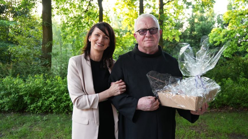Uśmiechnięta kobieta w jasnym żakiecie obejmuje ramię zakonnika w czarnym habicie, który trzyma w rękach zapakowany w celofan upominek. Oboje stoją w otoczeniu zieleni, na tle drzew i krzewów, w ciepłym świetle popołudnia.