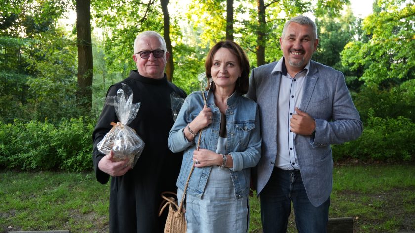 Dwóch mężczyzn i kobieta stoją razem na tle drzew, uśmiechając się do obiektywu, a jeden z nich trzyma zapakowany upominek.