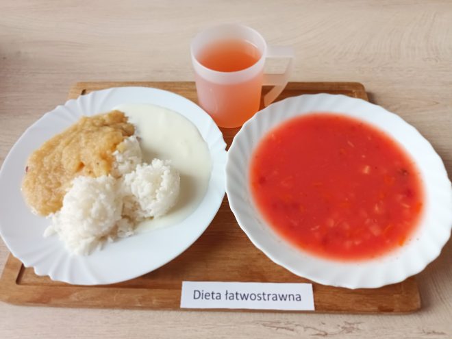 Zestaw obiadowy serwowany na białych talerzach i tacy drewnianej. Na pierwszym talerzu znajdują się trzy kulki białego ryżu polane jasnym sosem, z dodatkiem musu owocowego, prawdopodobnie z jabłek. Na drugim talerzu – zupa o czerwonym zabarwieniu, prawdopodobnie pomidorowa z warzywami. Do posiłku dołączono kubek z napojem, możliwe że kompotem. Na tacy znajduje się etykieta z opisem diety: „Dieta łatwostrawna”