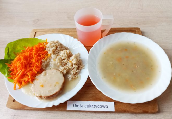 Na drewnianej tacy widoczne są trzy elementy składające się na posiłek oznaczony jako „Dieta cukrzycowa”. Po lewej stronie znajduje się talerz z drugim daniem: plaster pieczeni w jasnym sosie, porcja kaszy oraz starta na grubych oczkach marchewka podana na liściu sałaty. Obok, w głębokim talerzu, znajduje się jasna zupa jarzynowa z drobnymi kawałkami warzyw, głównie marchwi i ziemniaków. Uzupełnieniem jest różowy kompot w półprzezroczystym kubku z uchwytem. Całość została ustawiona schludnie na jasnym tle stołu
