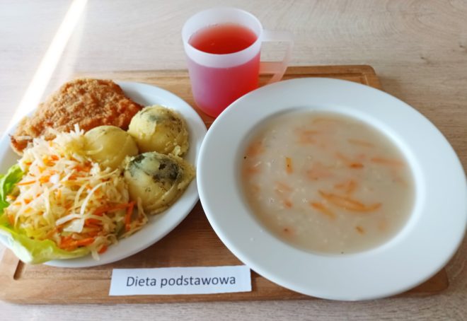 aca z obiadem w ramach diety podstawowej. Na pierwszym planie zupa marchwiowo-ryżowa w białym głębokim talerzu. Obok na oddzielnym talerzu: panierowany kotlet schabowy, trzy porcje ziemniaków (w tym jedna posypana koperkiem) oraz surówka z białej kapusty i marchewki na liściu sałaty. W tle plastikowy kubek z różowym kompotem. Całość ustawiona na tacy z etykietą „Dieta podstawowa”