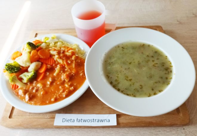 Zestaw obiadowy w ramach diety łatwostrawnej podany na drewnianej tacy. Na środku kartka z nadrukiem „Dieta łatwostrawna”. Po lewej stronie biały talerz z porcją makaronu świderki, polanego sosem mięsno-pomidorowym, oraz z dodatkiem gotowanych warzyw: marchewki, kalafiora i brokuła. Po prawej stronie głęboki biały talerz z zupą jarzynową z posiekaną natką pietruszki. W górnej części tacy półprzezroczysty kubek z czerwoną herbatką lub kompotem owocowym.