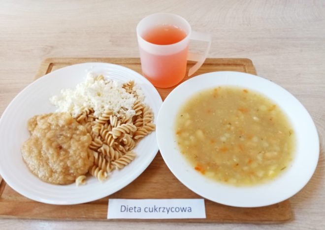 Taca z podpisem „Dieta cukrzycowa”. Na niej obiad zawierający talerz z zupą jarzynową (na bazie ziemniaków, marchwi, pietruszki), talerz z porcją pełnoziarnistego makaronu świderki, gotowanym kotletem mielonym i białym serem twarogowym, a także kubek z różowym kompotem lub naparem owocowym