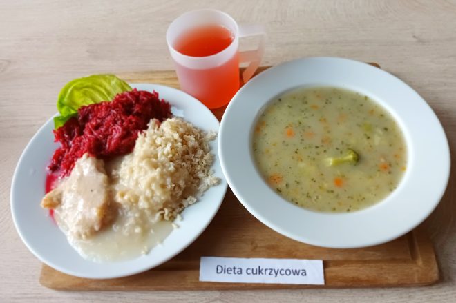 Zestaw obiadowy w diecie cukrzycowej: talerz z gotowanym kurczakiem w jasnym sosie, kaszą jęczmienną, buraczkami oraz plasterkiem sałaty i buraka. Obok miska z zupą jarzynową z brokułem, marchwią i ziemniakami. Do picia różowy napój w przezroczystym kubku. Na tacy kartka z napisem „Dieta cukrzycowa”