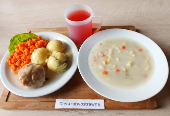 Obiad w diecie łatwostrawnej: porcja ziemniaków z koperkiem, gotowana marchewka, pulpet w jasnym sosie oraz liść sałaty. Obok zupa z makaronem świderki i warzywami (marchew, pietruszka). Do picia – czerwony napój w plastikowym kubku. Na tacy kartka z napisem „Dieta łatwostrawna”