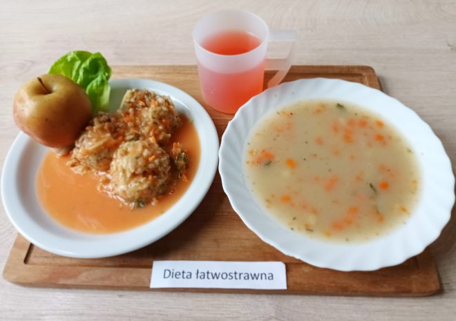 Taca z obiadem w diecie łatwostrawnej. W skład posiłku wchodzi zupa jarzynowa z drobno pokrojonymi warzywami, drugie danie z klopsami ryżowo-warzywnymi w sosie pomidorowym, liść sałaty oraz pieczone jabłko. Do picia różowy kompot w przezroczystym kubku z uchem