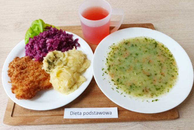 Taca z obiadem w diecie podstawowej. Danie składa się z zupy koperkowej z jarzynami, kotleta schabowego w panierce, ziemniaków z koperkiem oraz gotowanej czerwonej kapusty. Obok przezroczysty kubek z różowym napojem