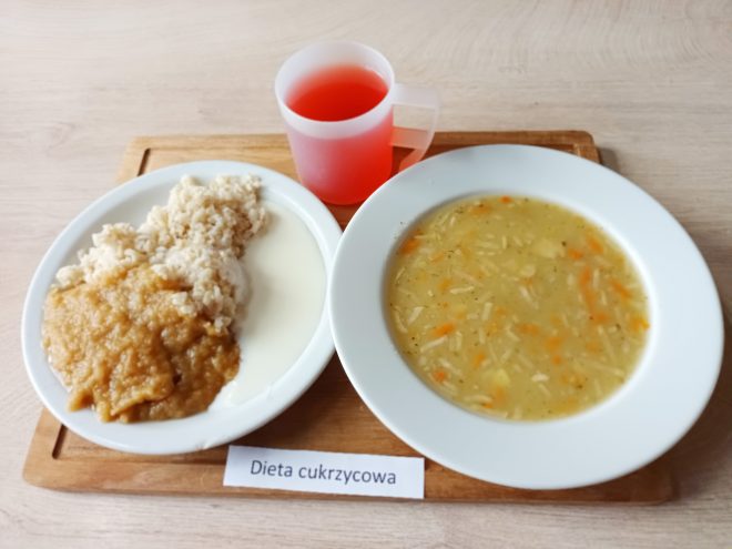 Taca z posiłkiem oznaczonym jako „Dieta cukrzycowa”. Na niej trzy elementy: talerz z ryżem polanym musem jabłkowym i mlekiem, talerz z zupą jarzynową z makaronem oraz przezroczysty kubek z czerwonym napojem. Wszystko podane na drewnianej podstawce