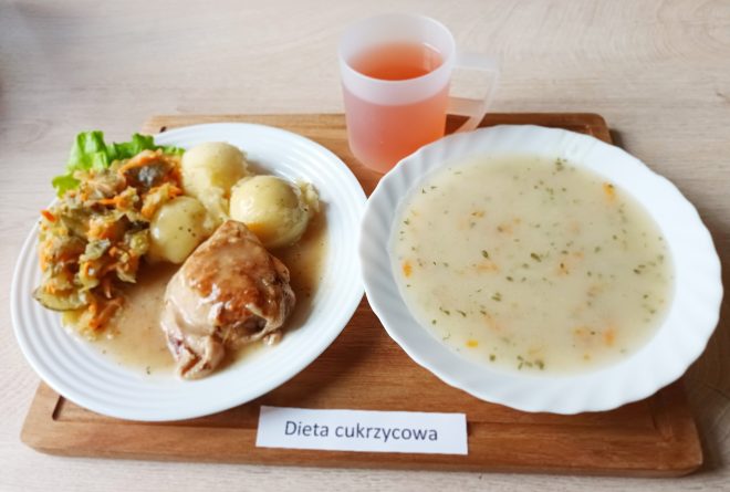 Taca z obiadem oznaczonym jako „Dieta cukrzycowa”. Na talerzu widoczne są ziemniaki z sosem, udko kurczaka w jasnym sosie oraz surówka z kiszonego ogórka i marchewki, ułożona na liściu sałaty. Obok znajduje się miska z zupą – najpewniej jarzynową na bazie wywaru warzywnego – i kubek z różowym napojem owocowym