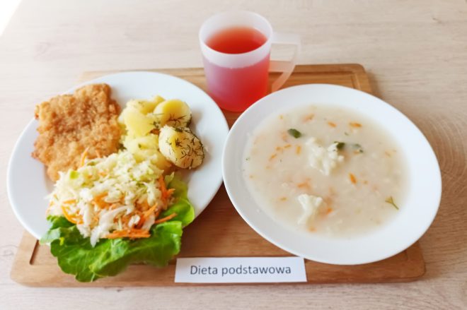 Zestaw obiadowy w diecie podstawowej serwowany na białym talerzu i drewnianej tacy. Składa się z panierowanego kotleta schabowego, gotowanych ziemniaków z koperkiem, surówki z białej kapusty i marchewki na liściu sałaty. Obok w białej misce zupa jarzynowa z kalafiorem, marchewką i ryżem. Do picia czerwony kompot w plastikowym kubku z uchem. Na tacy etykieta z napisem „Dieta podstawowa”