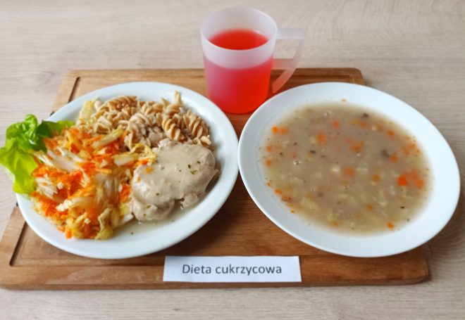Na zdjęciu przedstawiono zestaw obiadowy dla osób z cukrzycą. Na białym talerzu znajduje się gotowany filet drobiowy w lekkim sosie ziołowym, porcja pełnoziarnistego makaronu świderki oraz surówka z kapusty pekińskiej i tartej marchwi, udekorowana liściem sałaty. Obok znajduje się głęboki talerz z zupą jarzynową – widoczne są w niej kawałki marchwi, ziemniaków i przyprawy ziołowe. Całość uzupełnia czerwony napój podany w półprzezroczystym plastikowym kubku z uchem. Zestaw umieszczony jest na drewnianej tacce z etykietą „Dieta cukrzycowa”