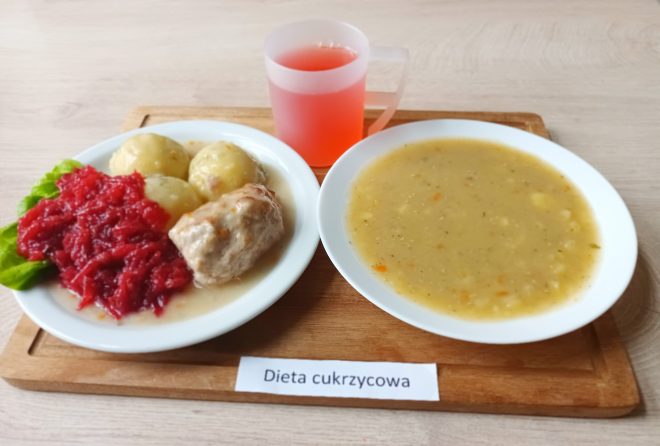 Obiad dedykowany osobom z cukrzycą. Na talerzu widać gotowane ziemniaki, pulpeta w sosie oraz duszone buraczki, ułożone na liściu sałaty. Obok znajduje się zupa krem – najpewniej z soczewicy lub grochu – z wyczuwalnymi kawałkami warzyw korzeniowych. Do obiadu podano czerwony napój w plastikowym kubku z uchem. Wszystko ustawione na drewnianej tacy z wyraźnym oznaczeniem „Dieta cukrzycowa”