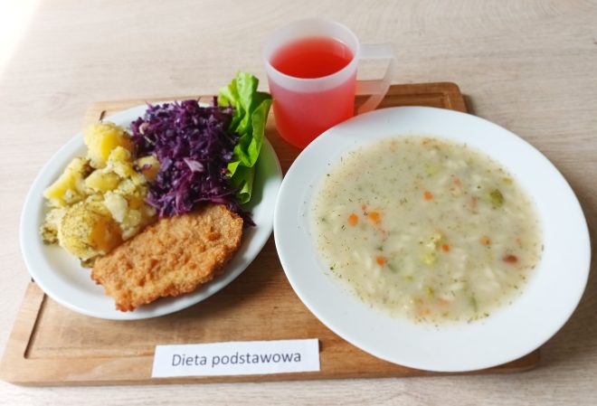 Taca z obiadem w diecie podstawowej: talerz z panierowanym kotletem, gotowanymi ziemniakami z koperkiem, surówką z czerwonej kapusty i listkami sałaty. Obok talerz z zupą jarzynową na białym sosie oraz kubek z czerwonym kompotem. Wszystko ustawione na drewnianej tacy z etykietą „Dieta podstawowa”