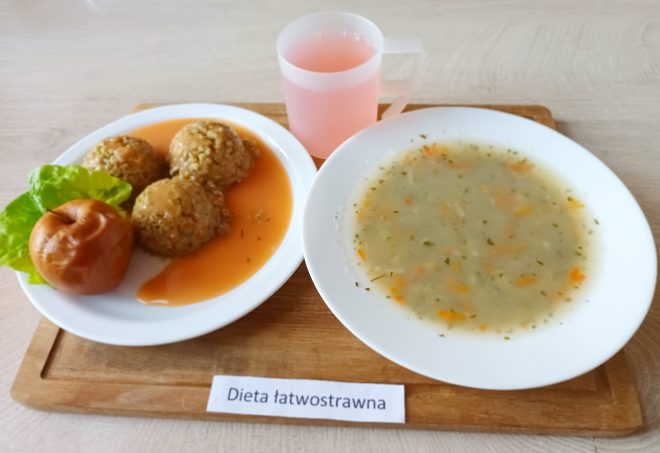 Obiad w diecie łatwostrawnej: talerz z trzema kulkami kaszy gryczanej w jasnym sosie, upieczone jabłko oraz liść sałaty. Obok talerz z klarowną zupą jarzynową oraz kubek z różowym napojem. Wszystko ustawione na drewnianej tacy z etykietą „Dieta łatwostrawna”