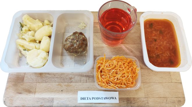 Obiad oznaczony jako „Dieta podstawowa”, ułożony na tacy z jasnego drewna. W trójdzielnym pojemniku znajdują się gotowane ziemniaki z koperkiem, pieczony kotlet mielony oraz miseczka czerwonego barszczu z warzywami i makaronem ryżowym. Obok – plastikowy kubek z czerwonym napojem oraz osobna przezroczysta miseczka z tartą marchewką. Danie kompletne i sycące, odpowiadające założeniom standardowej diety szpitalnej