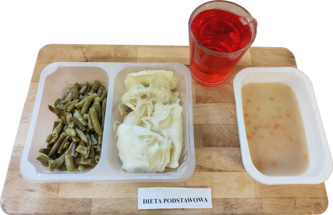 Na drewnianej tacy widnieje obiad w ramach „Diety podstawowej”. W trójdzielnej plastikowej tacce ułożono z lewej strony zieloną fasolkę szparagową, pośrodku – pierogi z kapustą i cebulką duszoną na parze, a z prawej strony – zupę jarzynową z drobno pokrojoną marchewką i ziemniakami. Towarzyszy im napój o intensywnie czerwonym kolorze, prawdopodobnie kompot z porzeczek lub wiśni, podany w przezroczystym kubku z uchem. Posiłek wygląda na lekki i zbilansowany