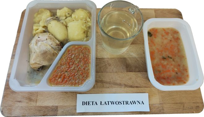 Zestaw obiadowy oznaczony jako „Dieta łatwostrawna”. Na tacy znajduje się pojemnik z trzema przegródkami: gotowane ziemniaki, gotowane udko kurczaka w lekkim sosie oraz duszona marchewka z cebulą. Obok pojemnika stoi kubek z klarownym napojem (najprawdopodobniej kompotem lub herbatą) oraz osobna miseczka z zupą jarzynową – widoczne kawałki marchwi i pietruszki. Całość ustawiona na desce z podpisem diety
