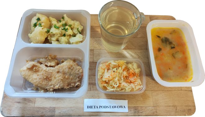Taca z obiadem w ramach diety podstawowej. W plastikowym naczyniu trzykomorowym: ugotowane ziemniaki z natką pietruszki, panierowany filet z kurczaka oraz porcja zupy ogórkowej w oddzielnym pojemniku. Obok szklanka z napojem w jasnym kolorze (herbata lub kompot) i mały pojemniczek z surówką z kiszonej kapusty i marchewki. Na tacy widoczna etykieta „DIETA PODSTAWOWA”.