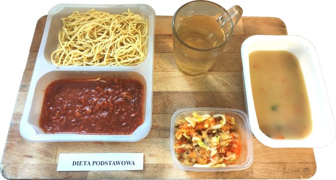 Taca z podpisem „DIETA PODSTAWOWA”. Na niej plastikowy pojemnik z dwoma przegródkami: w jednej znajduje się ugotowany makaron spaghetti, w drugiej – sos mięsny typu bolońskiego. Obok szklanka z jasnym naparem (herbata lub kompot), miska z jasną zupą jarzynową oraz pojemniczek z surówką z kiszonej kapusty, marchwi i cebuli
