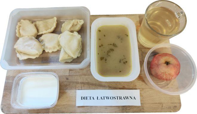 Taca z obiadem w diecie łatwostrawnej. Widać porcję pierogów z białym nadzieniem w przezroczystym pojemniku, zupę jarzynową w oddzielnym pojemniku, kubek z napojem (prawdopodobnie kompotem), obrane jabłko w plastikowym naczyniu oraz kubeczek z jogurtem lub kefirem. Na tacy znajduje się karteczka z napisem „DIETA ŁATWOSTRAWNA”