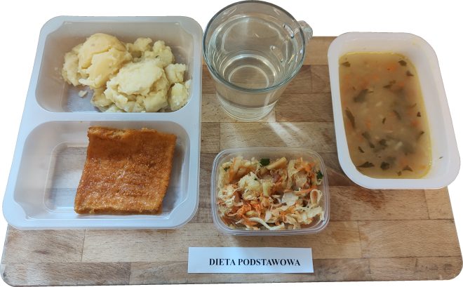 Taca z obiadem w ramach diety podstawowej. Na niej: porcja gotowanych ziemniaków, panierowany i smażony filet rybny, surówka z kapusty kiszonej z marchewką i pietruszką, zupa jarzynowa z natką pietruszki w oddzielnym pojemniku oraz kubek z przegotowaną wodą. Przed tacką znajduje się etykieta z napisem: „DIETA PODSTAWOWA”