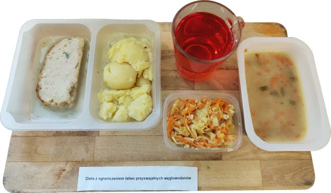 Taca z obiadem szpitalnym przeznaczonym dla pacjentów na diecie z ograniczeniem łatwo przyswajalnych węglowodanów. Na tacy znajdują się: Porcja gotowanych ziemniaków, Kawałek pieczeni drobiowej lub pulpetu pieczonego w lekkim sosie, Surówka z marchwi i kapusty, Zupa jarzynowa o jasnej barwie z kawałkami warzyw, Czerwony napój owocowy w przezroczystym kubku. Wszystko podane w plastikowych pojemnikach na drewnianej desce z opisem diety