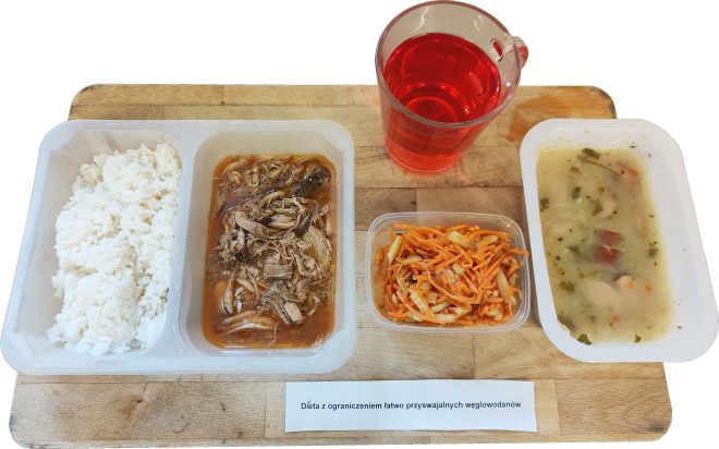 Taca obiadowa z dietą niskowęglowodanową. Na tackach: gotowany biały ryż, szarpane mięso (prawdopodobnie wołowina lub wieprzowina) w sosie, surówka z marchewki i selera, zupa jarzynowa z ziemniakami i natką pietruszki oraz czerwony napój w przezroczystym kubku