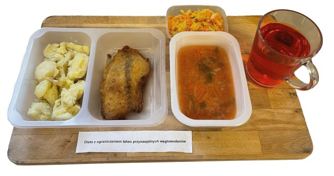 Zestaw obiadowy składający się z gotowanych ziemniaków, panierowanego filetu rybnego, czerwonego barszczu z warzywami, surówki z kapusty i marchwi oraz czerwonego napoju w szklance. Na tacy etykieta: „Dieta z ograniczeniem łatwo przyswajalnych węglowodanów”