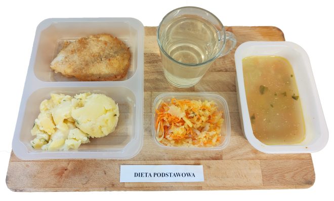 Obiad w diecie podstawowej zapakowany w pojemniki: ziemniaki z koperkiem, panierowany kotlet schabowy oraz surówka z kapusty pekińskiej, marchewki i jabłka. Obok osobny pojemnik z zupą jarzynową oraz szklanka klarownego napoju. Całość ułożona na drewnianej tacy z etykietą „DIETA PODSTAWOWA”
