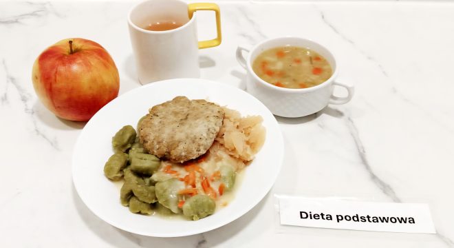 Na zdjęciu znajduje się zestaw obiadowy w diecie podstawowej. Na białym talerzu ułożono kotlet mielony, porcję zielonych klusek polanych sosem z marchewką oraz duszoną kapustę. Obok znajduje się czerwone jabłko, kubek z herbatą oraz miseczka z zupą jarzynową z widocznymi kawałkami marchwi i ziemniaków. Na stole leży etykieta z napisem "Dieta podstawowa"