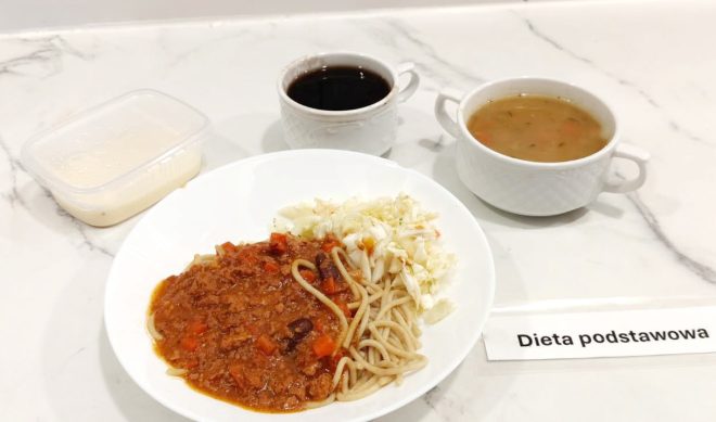 Talerz z makaronem spaghetti z sosem bolońskim i surówką z kapusty. Obok filiżanka ciemnego napoju, zupa jarzynowa w białej miseczce oraz deser mleczny w plastikowym pojemniku. Na stoliku etykieta z napisem „Dieta podstawowa”