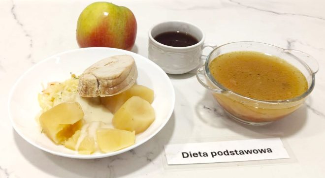 Talerz z gotowanym filetem z kurczaka, gotowanymi ziemniakami i surówką z białej kapusty. Obok filiżanka ciemnego napoju, miseczka z bulionem warzywnym oraz czerwone jabłko. Na stoliku etykieta z napisem „Dieta podstawowa”