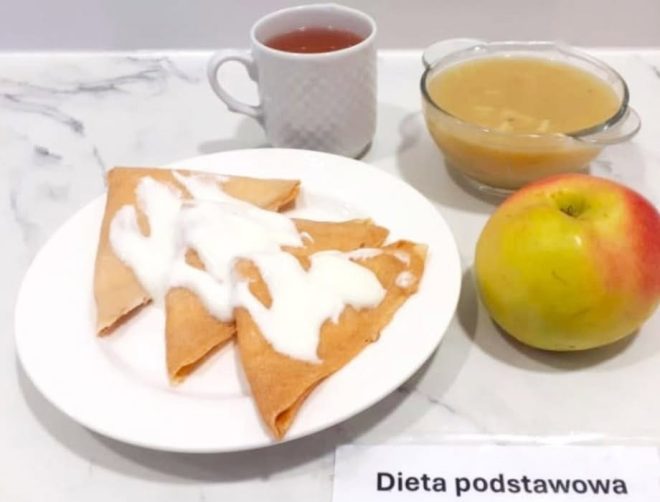 Zestaw obiadowy z naleśnikami z nadzieniem owocowym, polanymi jogurtem naturalnym, podany z jabłkiem, kompotem owocowym i filiżanką czerwonej herbaty. Na stoliku widoczna etykieta ‘Dieta podstawowa