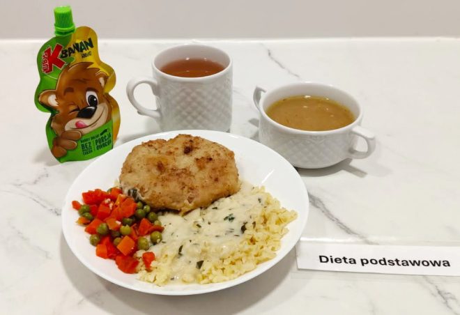 Taca z etykietą „Dieta podstawowa”. Na niej znajduje się talerz z panierowanym kotletem, drobnym makaronem w sosie śmietanowo-ziołowym oraz gotowanymi warzywami (marchewka i zielony groszek). Obok dwa ceramiczne kubki – jeden z jasną zupą, drugi z ciemniejszym naparem (herbatą). Dodatkowo saszetka przecieru owocowego typu „Kubuś” dla dzieci