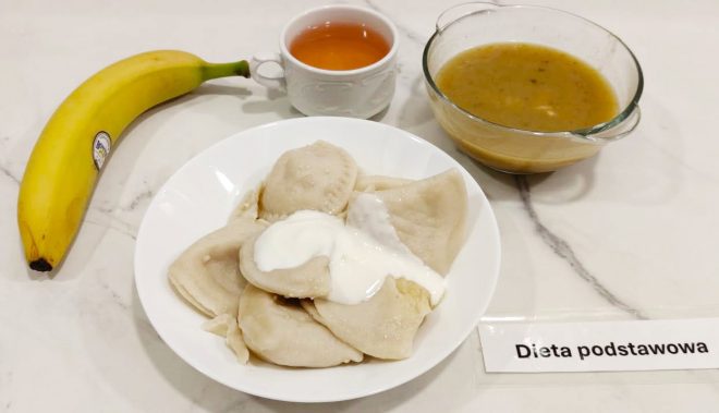 Zestaw obiadowy w diecie podstawowej. Na marmurowym blacie ustawiono biały talerz z porcją pierogów polanych śmietaną. Towarzyszy im szklana miska z zupą (najprawdopodobniej jarzynową lub grochową) o złocistobrązowym kolorze. Po lewej stronie znajduje się dojrzały banan oraz filiżanka z herbatą. Przed talerzem widoczna jest karteczka z napisem „Dieta podstawowa”