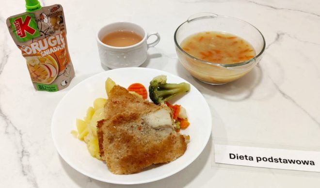 Obiad w diecie podstawowej: panierowany filet rybny, gotowane ziemniaki oraz mieszanka warzyw gotowanych na parze (brokuł, marchewka, kalafior). Z boku miseczka z zupą jarzynową oraz kubek herbaty. W tle opakowanie musu owocowego „Kubuś Drugie Śniadanie”