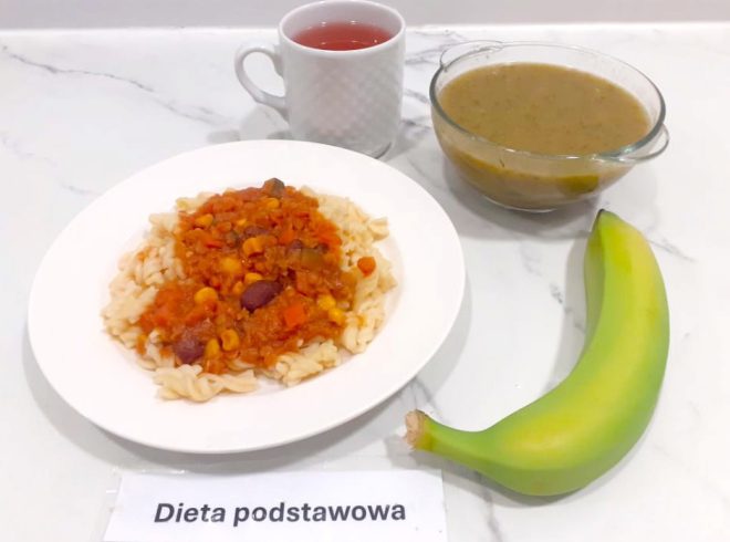Obiad w diecie podstawowej: makaron świderki z warzywnym sosem meksykańskim, obok miseczka z zupą jarzynową i cały banan. Do picia czerwony napar w białym kubku. Posiłki ustawione na jasnym blacie. Etykieta z napisem „Dieta podstawowa”