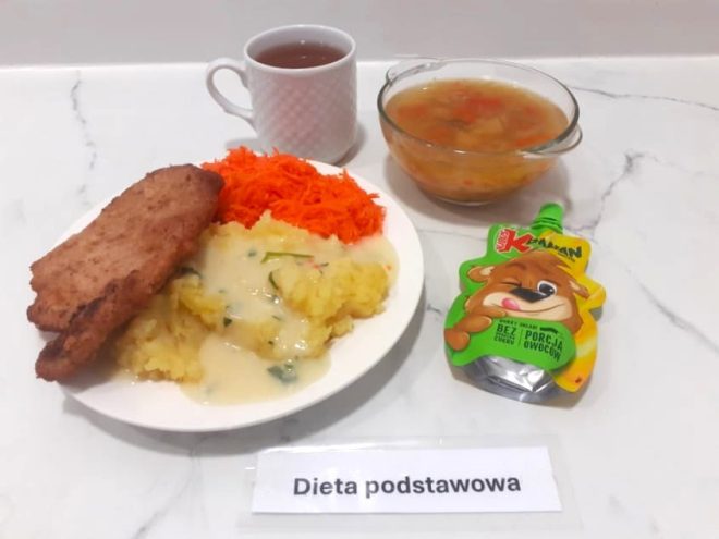 Talerz z panierowanym kotletem, ziemniakami z sosem i tartą marchewką. Obok miseczka zupy jarzynowej, herbata oraz mus owocowy w tubce z grafiką misia. Dieta podstawowa